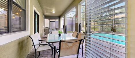 A covered patio area with a glass table and chairs, a view of a swimming pool, and a door leading to an outdoor space.
