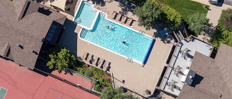 Aerial view of the property featuring a sparkling outdoor swimming pool surrounded by lounge chairs, tennis court, and landscaped green spaces with ample parking nearby.