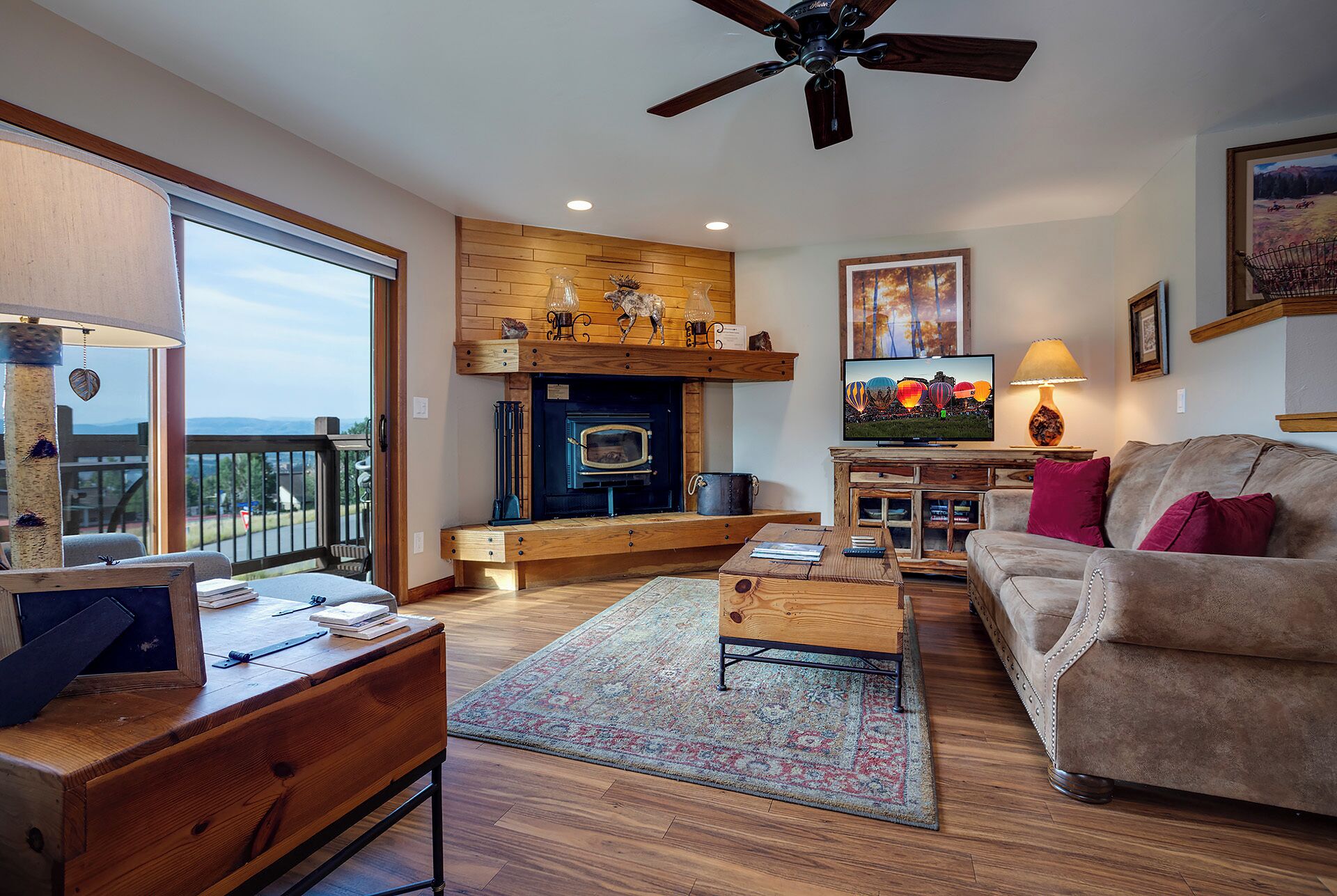 A cozy living room with a fireplace, wooden furniture, and a large window overlooking a scenic view.
