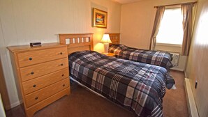 Une chambre avec deux lits, une commode en bois, une table de chevet avec une lampe, un tableau encadré au mur et une fenêtre avec des rideaux.