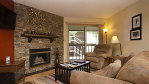 A living room with a stone fireplace, a comfortable sofa, a chair, a coffee table, and a view through a glass door to an outdoor balcony. - SHB39 | Nice Mtn Condo | Hot Tub | On Free City Bus Route (Steamboat Springs)