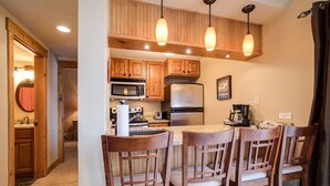 Una cocina con armarios de madera, un refrigerador de acero inoxidable, un microondas y una cafetera. Hay cuatro sillas de madera con asientos acolchados.