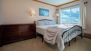 A bedroom with a bed, a wooden dresser, a painting, and a window with a view of a snowy landscape. - SM436 | Charming Condo | Mountain Views | Hot Tub | Ski In | Free City Bus (Steamboat Springs)