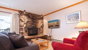 A living room with a stone fireplace, a television mounted on the wall, a red armchair, and a wooden coffee table. - SM436 | Charming Condo | Mountain Views | Hot Tub | Ski In | Free City Bus (Steamboat Springs)