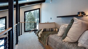 A bedroom with a bed, a window with a view of trees, a wooden staircase, and a ceiling with exposed beams.