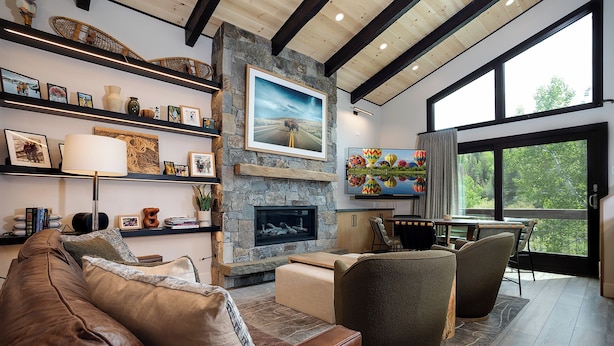 A modern living room with a stone fireplace, leather sofa, and a dining area with a large window offering a view of the outdoors.