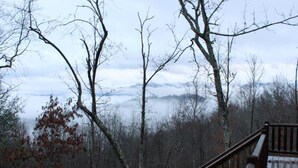 A wooden deck with a railing overlooking a forested area with bare trees and a distant mountain range. - Sunrise Ridge - Free Train Tours + Cabin + Stunning Views/Deck + Close to (Bryson City)