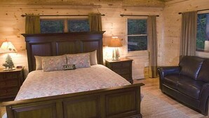 A log cabin bedroom with a wooden bed, a leather sofa, and a nightstand with a lamp. - Sunrise Ridge - Free Train Tours + Cabin + Stunning Views/Deck + Close to (Bryson City)