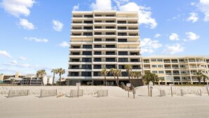 Un hotel a più piani con balconi, situato su una spiaggia sabbiosa con palme.