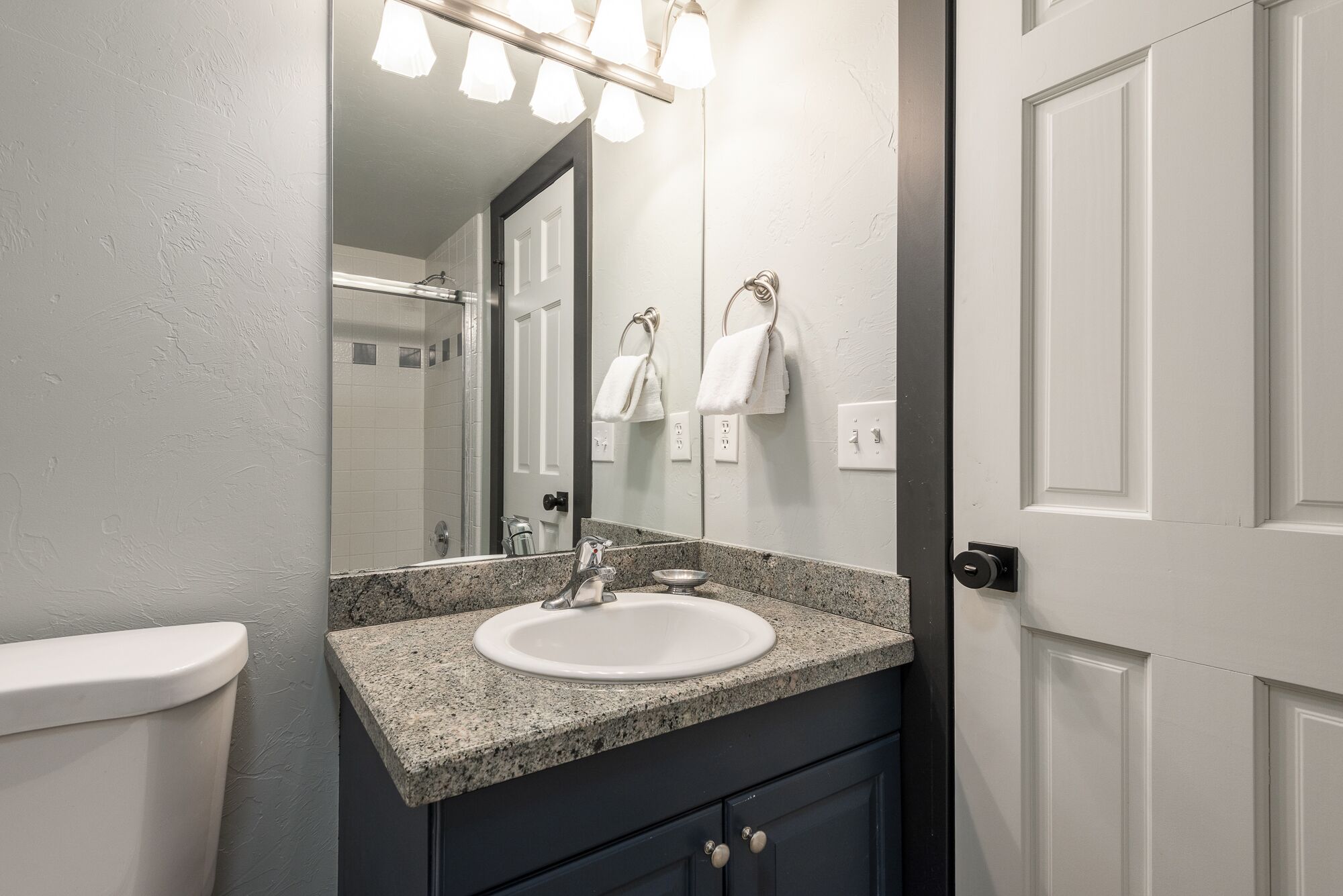 Un baño con encimera de granito, lavabo blanco y un espejo encima. Hay toallas colgadas en un perchero y una puerta a la derecha.