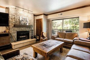 A living room with a stone fireplace, leather sofas, a wooden coffee table, and a large window with a view of trees.