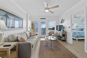 A well-lit living room with a sofa, coffee table, and a television. There is a ceiling fan, a door leading outside, and a bed in the adjacent room.
