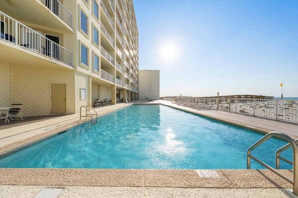 A swimming pool adjacent to a multi-story building with balconies, a clear sky, and a distant view of the ocean.
