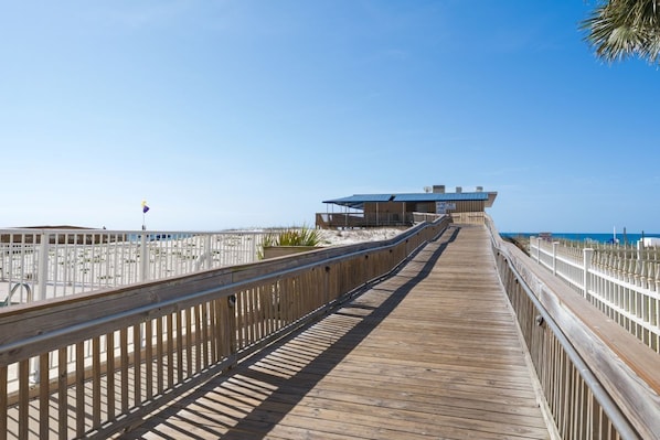 A wooden boardwalk leading to a building with a blue roof, white railings, and a clear sky. - Gulf Shores Beachfront Retreat | Sleeps 6 | Gulf-Front Pool & On-Site Dining (Gulf Shores)