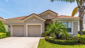Una casa a due piani con facciata beige, due porte del garage e un piccolo giardino con palme.