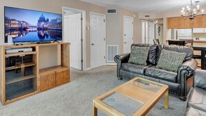 Una sala de estar con televisor de pantalla plana, sofá de cuero, mesa de centro de vidrio, mueble de entretenimiento de madera y una zona de cocina con gabinetes de madera y una araña.