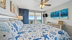 Ein Schlafzimmer mit einem Bett, ausgestattet mit einem blau-weißen, marine-inspirierten Bettbezug, einem Deckenventilator, einem großen Fenster mit Blick und Kunstwerken an den Wänden.