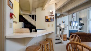 Une cuisine avec un comptoir blanc, des tabourets en bois et un escalier menant à un espace de vie comprenant un téléviseur et une cheminée.