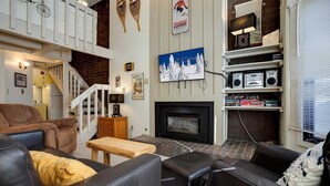 Un salon chaleureux avec une cheminée, un escalier et une collection de livres et d’objets décoratifs disposés sur des étagères.