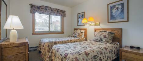 A hotel room with two beds, a window with blinds, a lamp, a clock, and two framed pictures on the wall.
