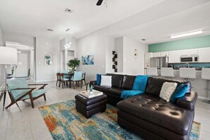 Uma sala de estar moderna com um sofá de couro escuro, uma área de jantar com mesa e cadeiras, e uma cozinha com armários e eletrodomésticos brancos.