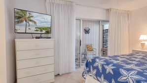 A hotel room with a bed, a TV on a dresser, a balcony with a chair, and a view of the beach.