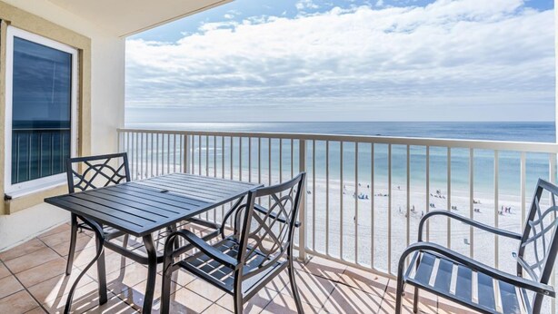 Een balkon met een tafel en stoelen, uitkijkend op een strand met mensen en een heldere lucht.