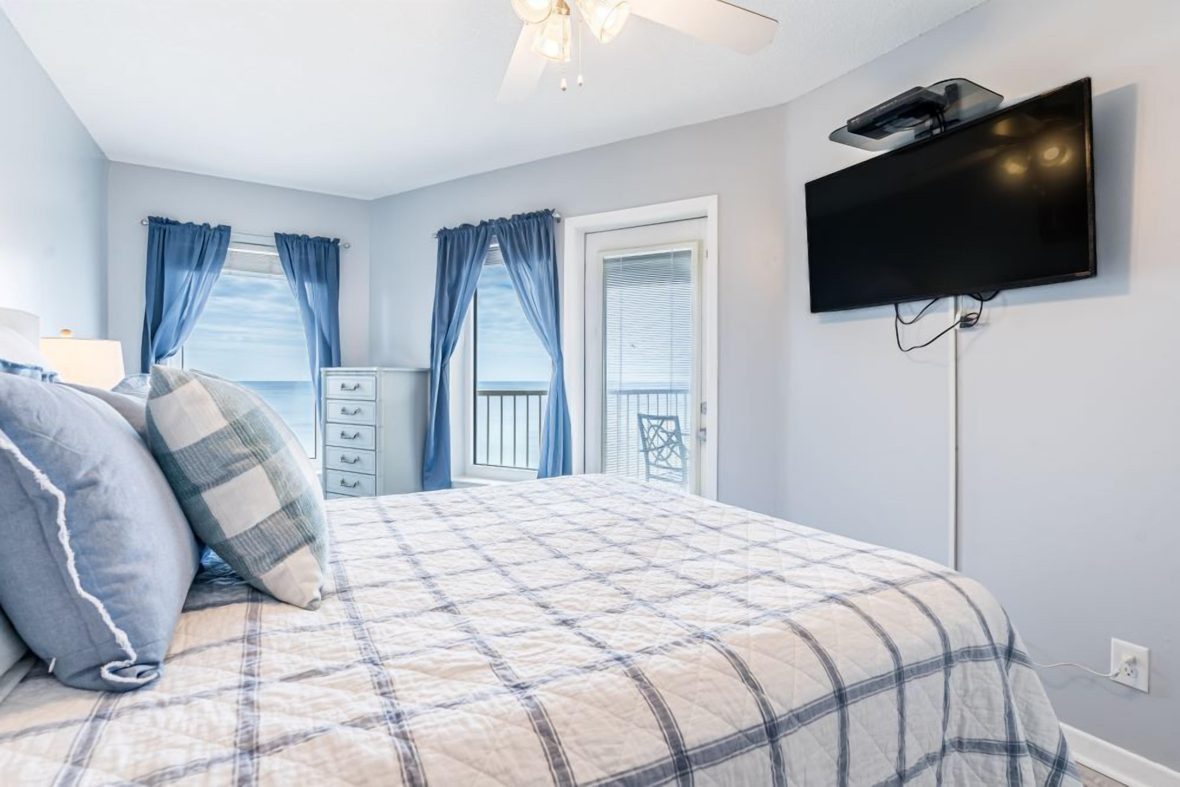 Een slaapkamer met een bed, een dressoir, een aan de muur bevestigde televisie en een balkon met blauwe gordijnen.