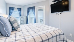 Een slaapkamer met een bed, een dressoir, een aan de muur bevestigde televisie en een balkon met blauwe gordijnen.