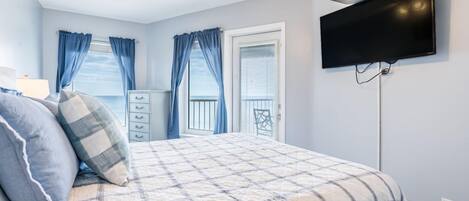Een slaapkamer met een bed, een dressoir, een aan de muur bevestigde televisie en een balkon met blauwe gordijnen.