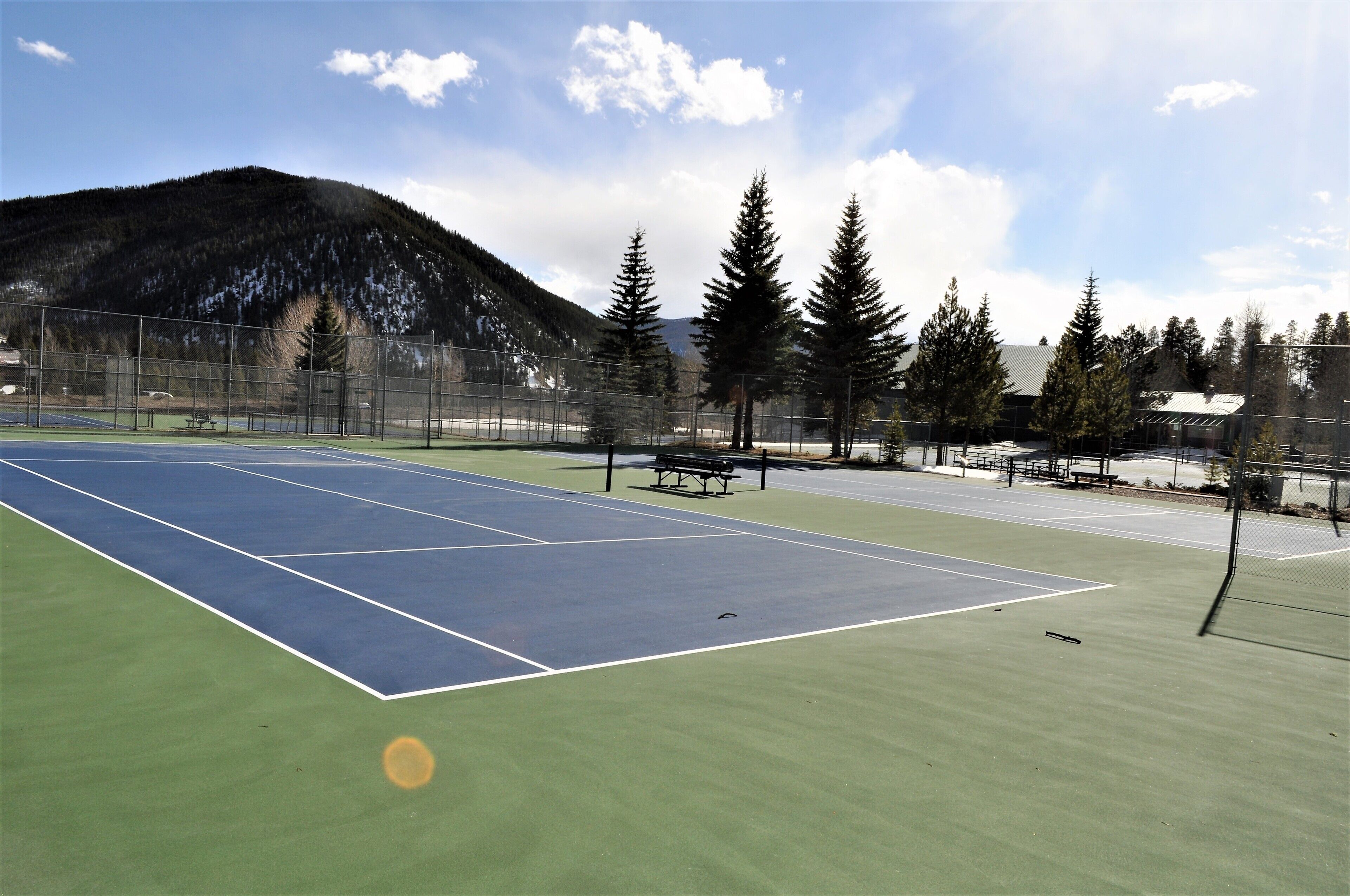 Un terrain de tennis avec un filet, entouré d’une clôture, de bancs et d’arbres.