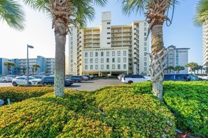 A multi-story hotel with a central entrance, surrounded by a parking lot and palm trees. - Beachfront Gulf Shores Getaway | Sleeps 6 | Resort Pool & On-Site Dining (Gulf Shores)