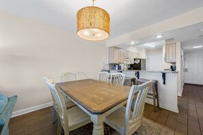 A dining area with a wooden table and chairs, a hanging light fixture, and a kitchen in the background. - Beachfront Gulf Shores Getaway | Sleeps 6 | Resort Pool & On-Site Dining (Gulf Shores)