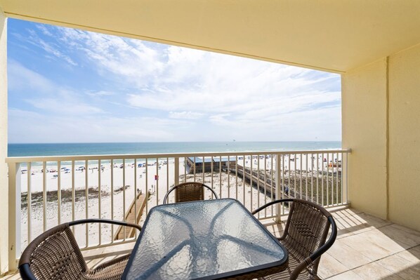 A balcony with a table and chairs, overlooking a beach with a fence and beachgoers. - Beachfront Gulf Shores Getaway | Sleeps 6 | Resort Pool & On-Site Dining (Gulf Shores)