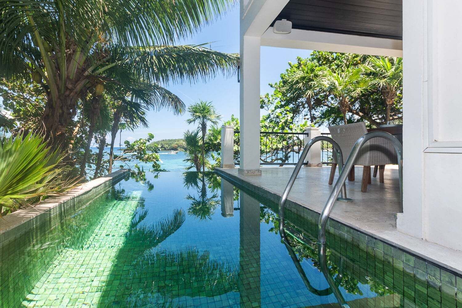 A clear pool with a view of the sea, surrounded by palm trees and a covered seating area.