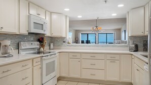 A modern kitchen with white cabinets, a stainless steel stove, and a large window overlooking the ocean. - Surf Dweller 504 by Alicia Hollis Rentals FREE ADVENTURES $300/Day (Fort Walton Beach)