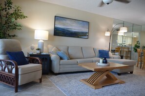 A living room with a sofa, armchair, coffee table, lamp, and a painting on the wall. - Surf Dweller 504 by Alicia Hollis Rentals FREE ADVENTURES $300/Day (Fort Walton Beach)