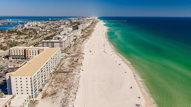 A coastal cityscape with a long sandy beach, buildings along the shore, and a clear blue sky. - Surf Dweller 504 by Alicia Hollis Rentals FREE ADVENTURES $300/Day (Fort Walton Beach)