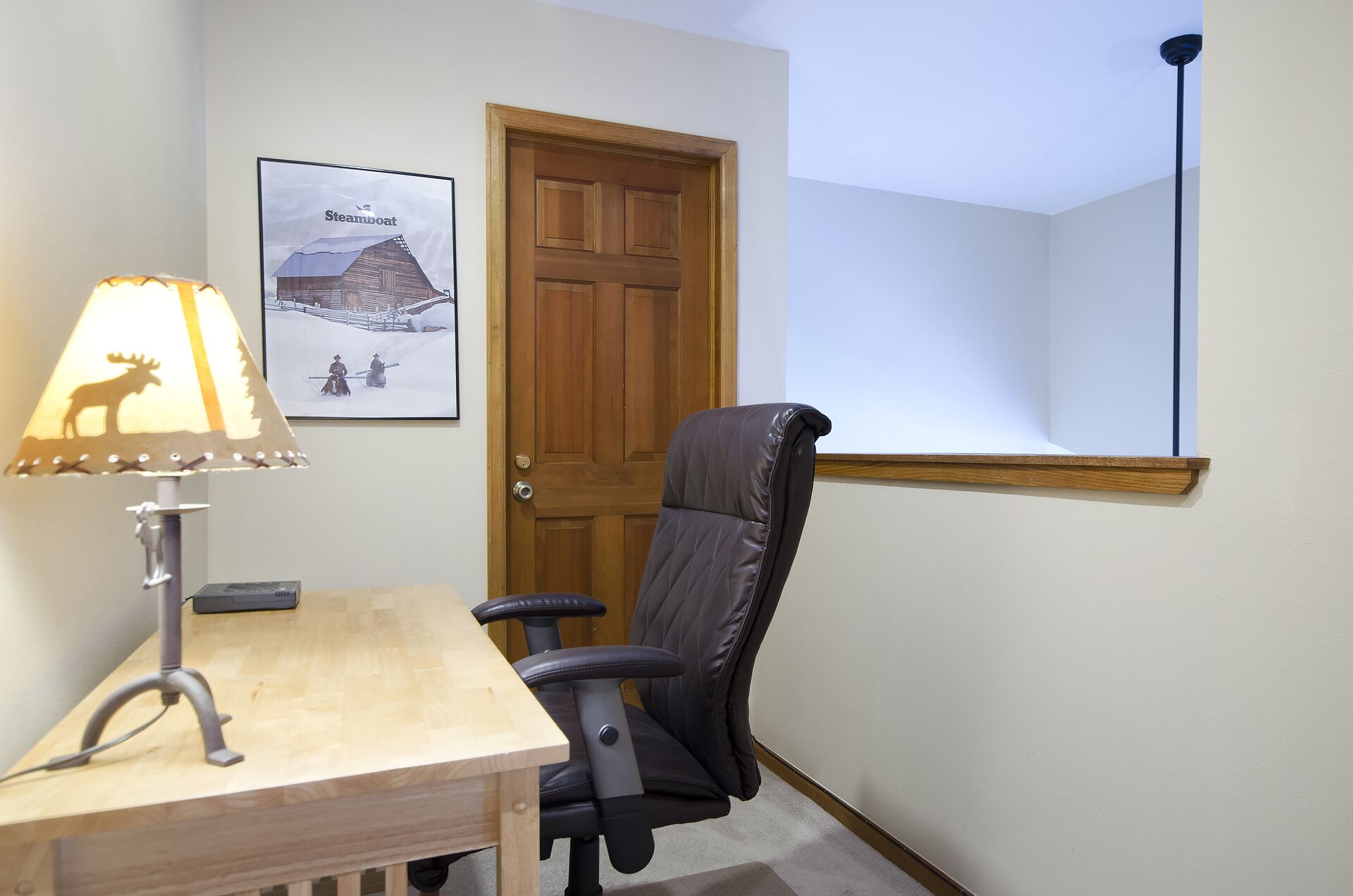 Une pièce avec un bureau en bois, un fauteuil de bureau noir, une lampe dont l’abat-jour est orné de la silhouette d’un élan, un tableau encadré représentant un paysage enneigé et une porte en bois.