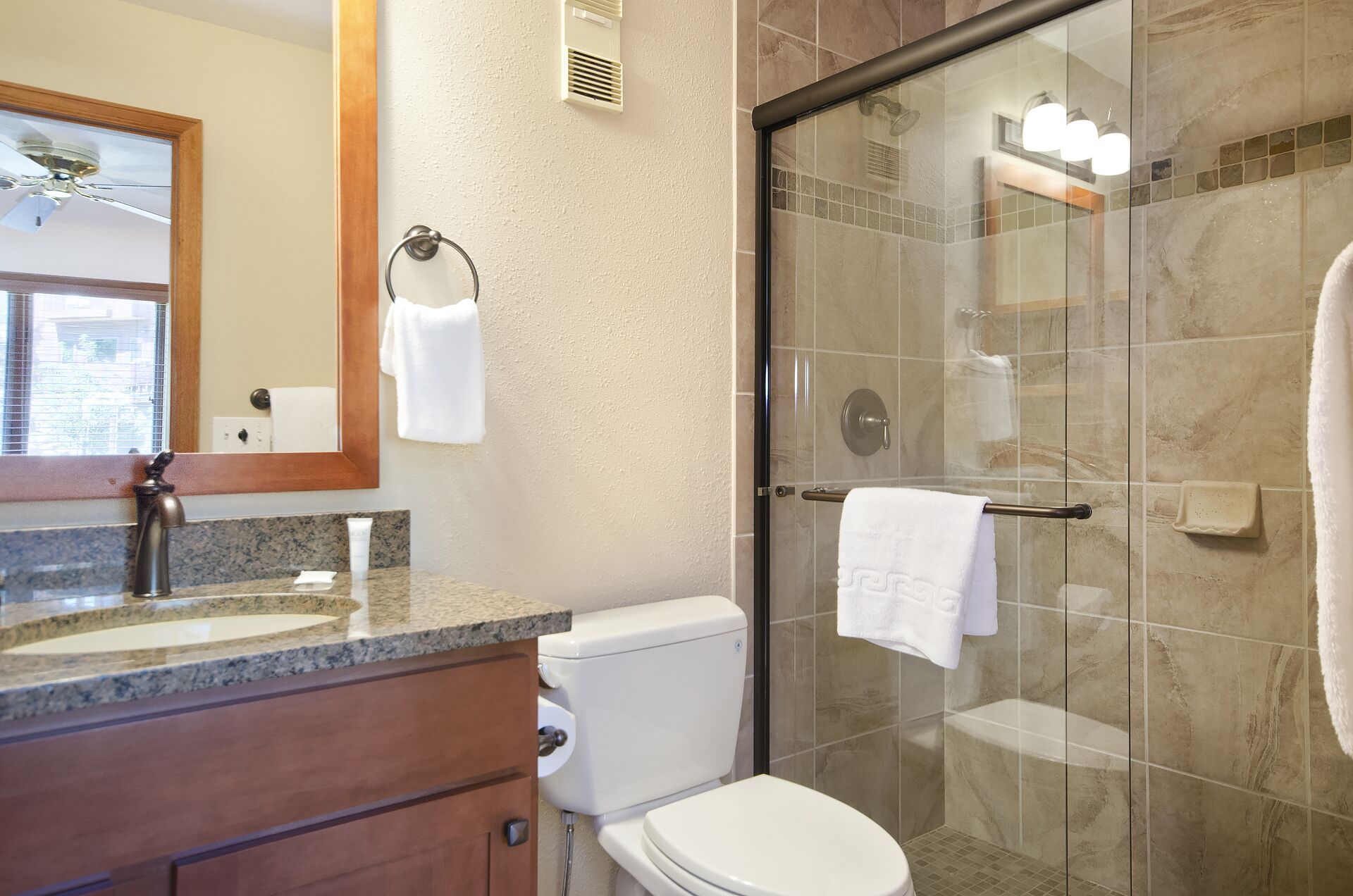 A bathroom with a granite countertop, a white toilet, a glass shower enclosure, and a mirror with a towel hanging on the wall.