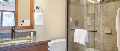 A bathroom with a granite countertop, a white toilet, a glass shower enclosure, and a mirror with a towel hanging on the wall.