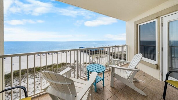 A balcony with white chairs and a blue table, overlooking a beach with a clear sky. - Spacious Beachfront Gulf Shores Condo | Sleeps 10 | Pool & On-Site Dining (Gulf Shores)