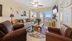 A living room with a ceiling fan, a sofa, a coffee table, and a TV mounted on the wall. - Spacious Beachfront Gulf Shores Condo | Sleeps 10 | Pool & On-Site Dining (Gulf Shores)
