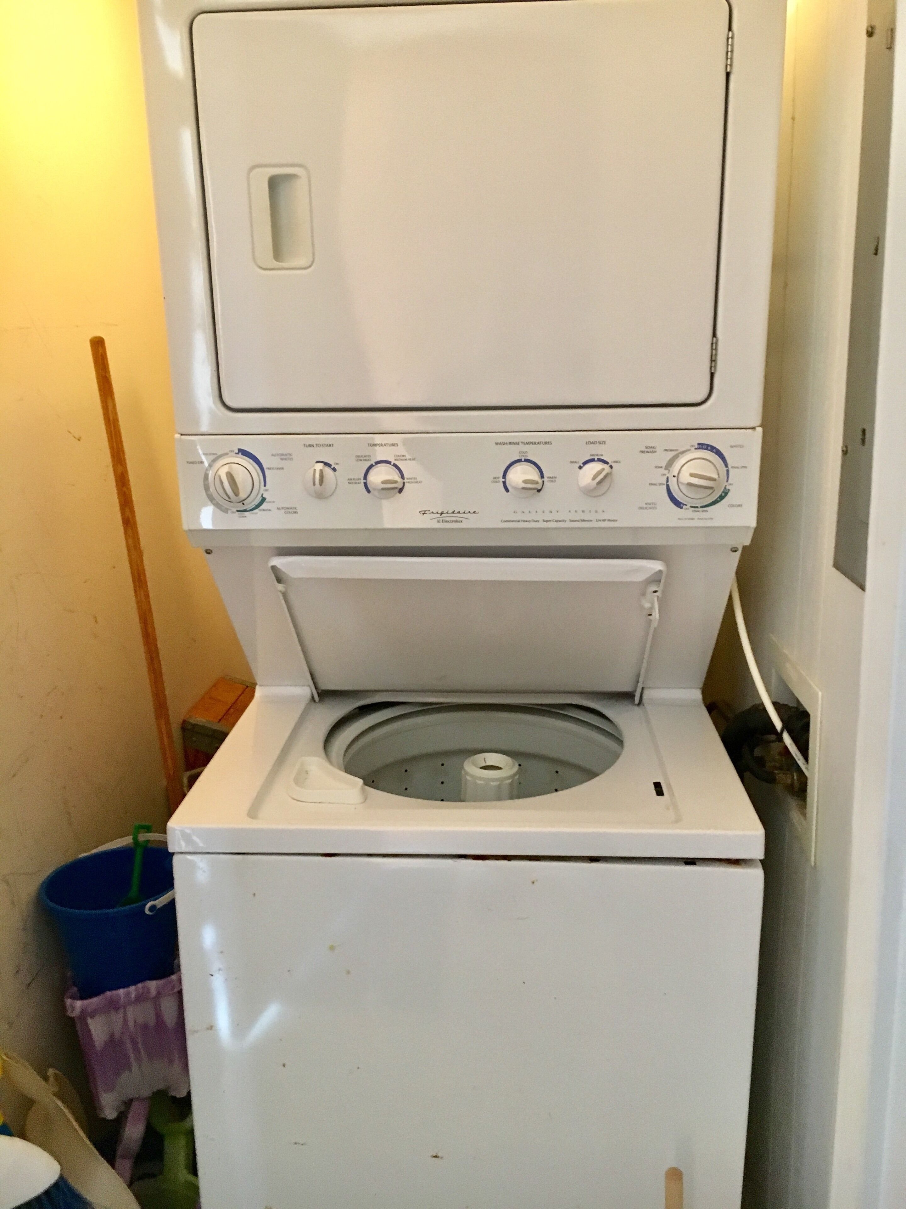 Une machine à laver à chargement frontal, dotée d’un panneau de commande, d’un seau bleu et d’un balai.