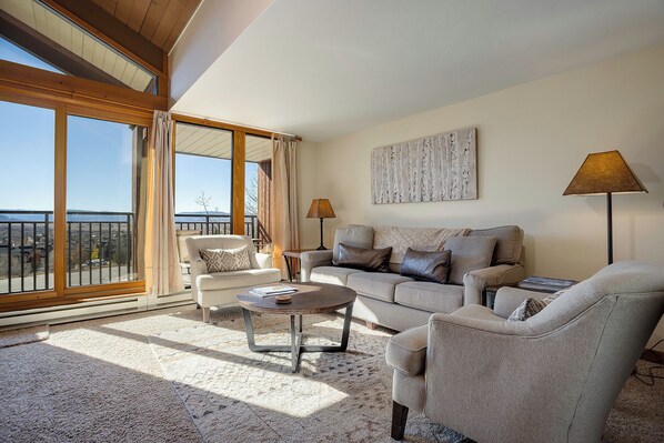 A spacious living room with a large window, a sofa set, a coffee table, and a painting on the wall.