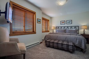 A bedroom with a bed, two bedside lamps, a chair, and two windows with blinds.