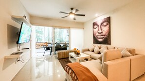 A modern living room with a sofa, coffee table, and TV. There is a ceiling fan and a large window with a view of trees.