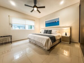 Quarto com cama, ventoinha no teto, mesa de cabeceira, janela e um quadro na parede.