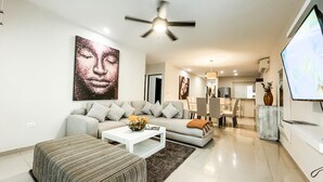 A modern living room with a sofa, ottoman, and coffee table, a dining area with chairs, and a TV on the right.