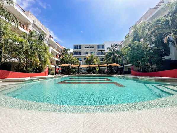 A clear swimming pool with lounge chairs and umbrellas, surrounded by palm trees and a multi-story building.